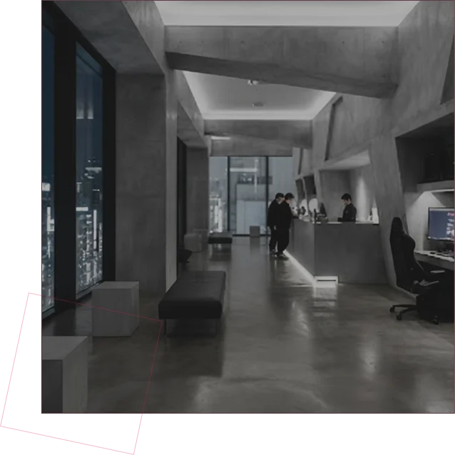 Interno di una sala da gaming moderna e futuristica con pareti in cemento, luci soffuse, ampie vetrate panoramiche con vista sulla città di notte e postazioni con computer.